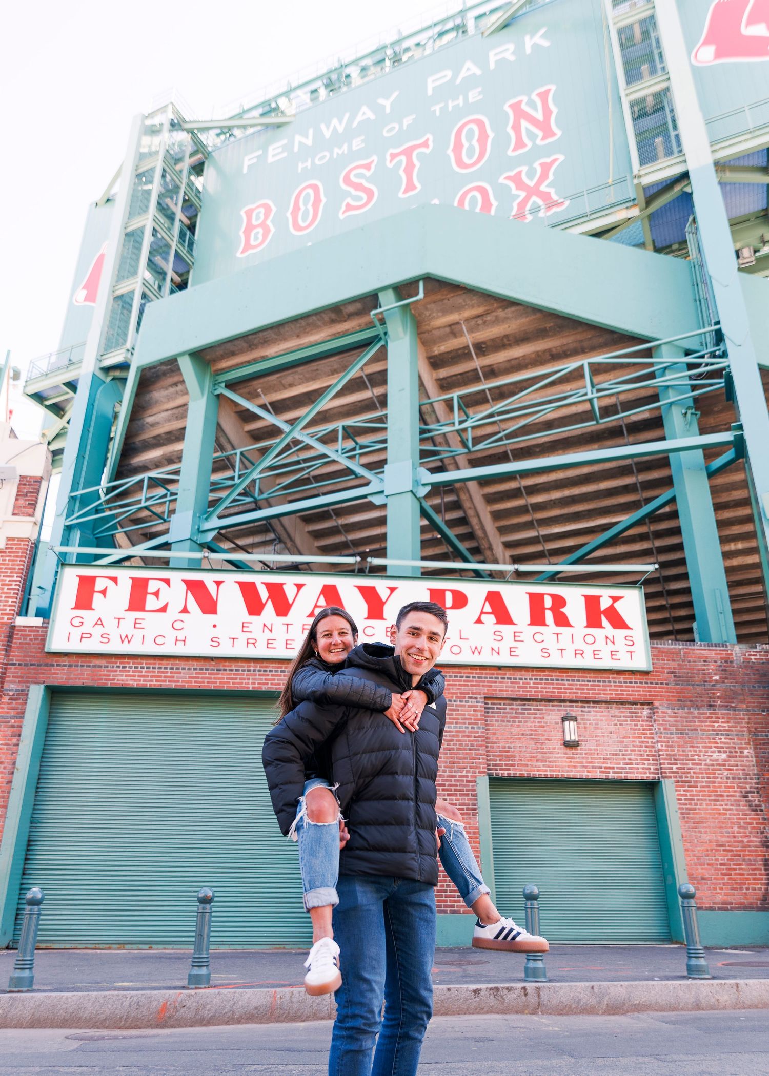 Couple posing playfully in front of Fenway Park, home of the Boston Red Sox, wearing winter jackets.