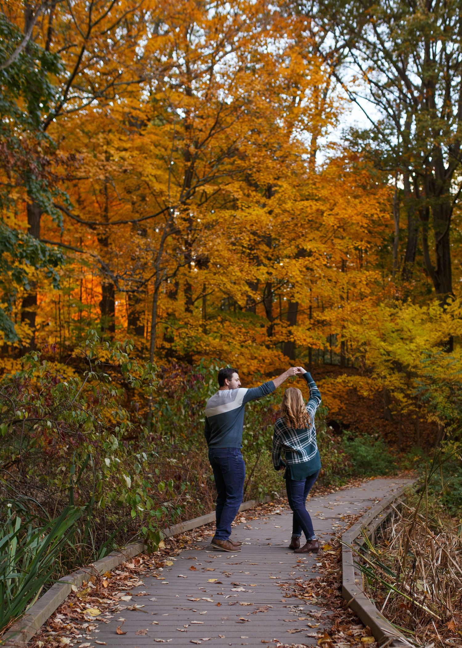 Couple dancing on a wooden path surrounded by vibrant orange and yellow autumn foliage in a scenic forest.