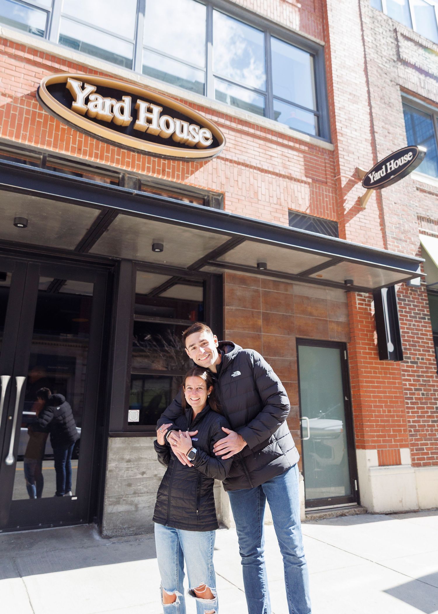 Couple posing and hugging in front of Yard House restaurant brick building entrance.
