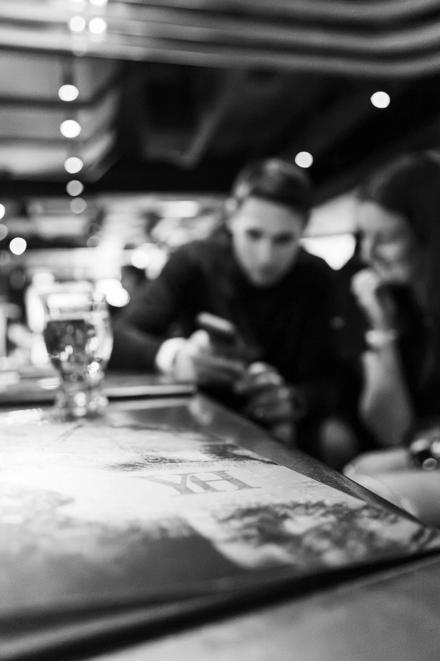 Black and white photo of two people at a bar table with a beer glass, one checking a phone.