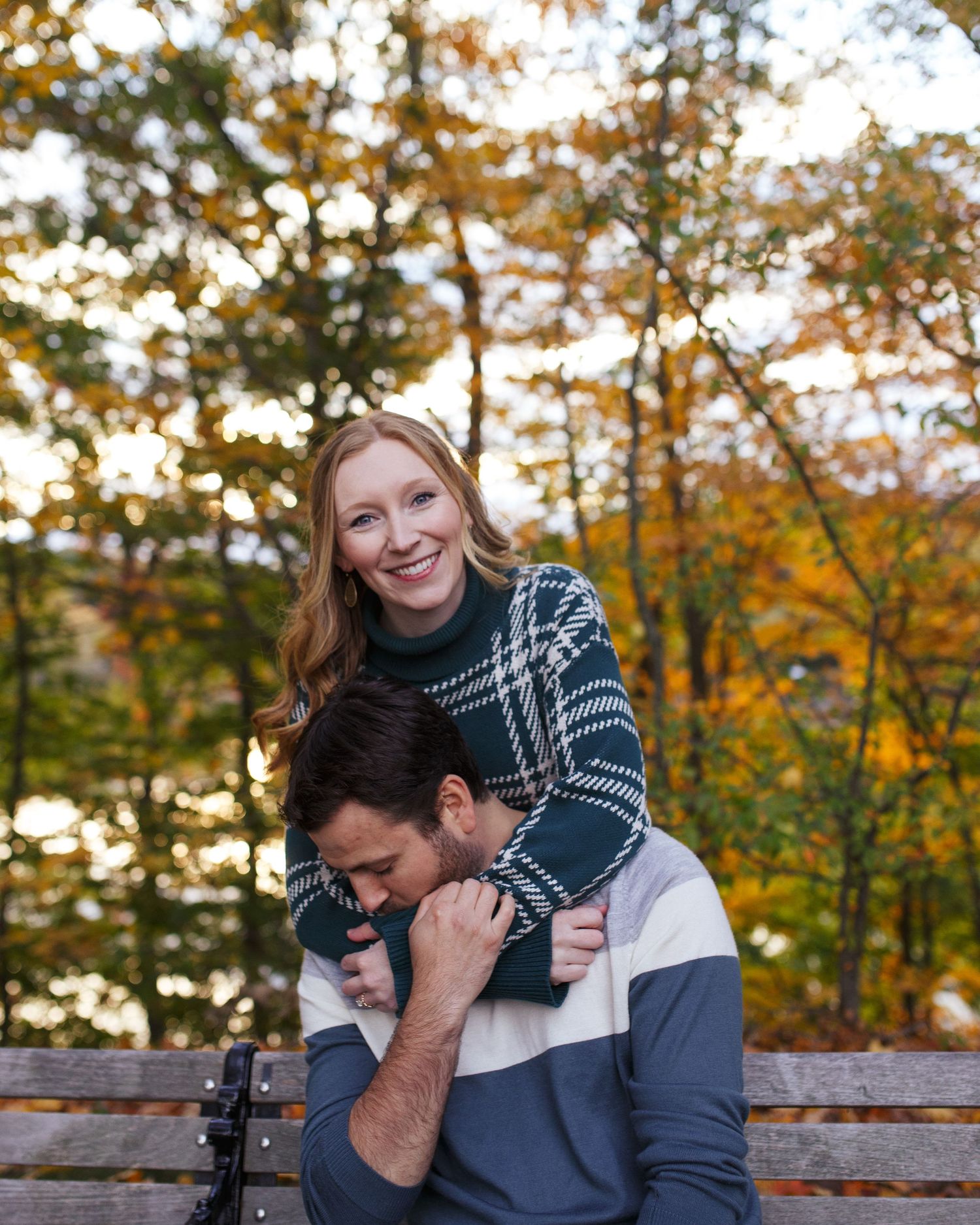 A couple shares a tender moment together on a bench during golden hour with autumn trees in the background.