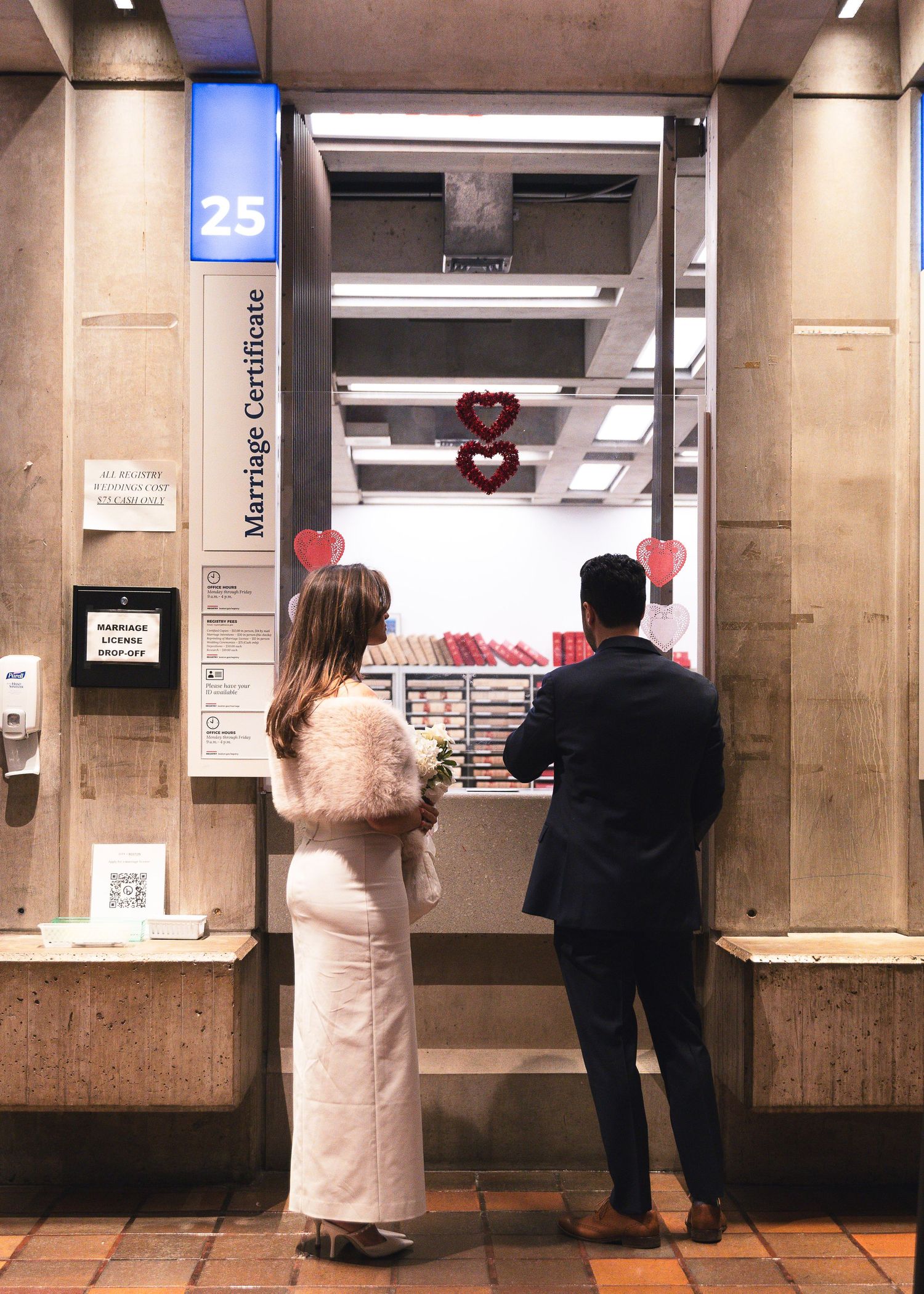 Couple in wedding attire obtaining marriage certificate at counter 25, decorated with red hearts.