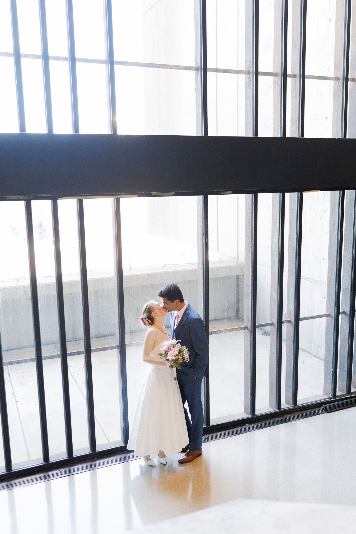 Bride and groom share a romantic kiss in a sunlit modern venue with floor-to-ceiling windows on their wedding day.