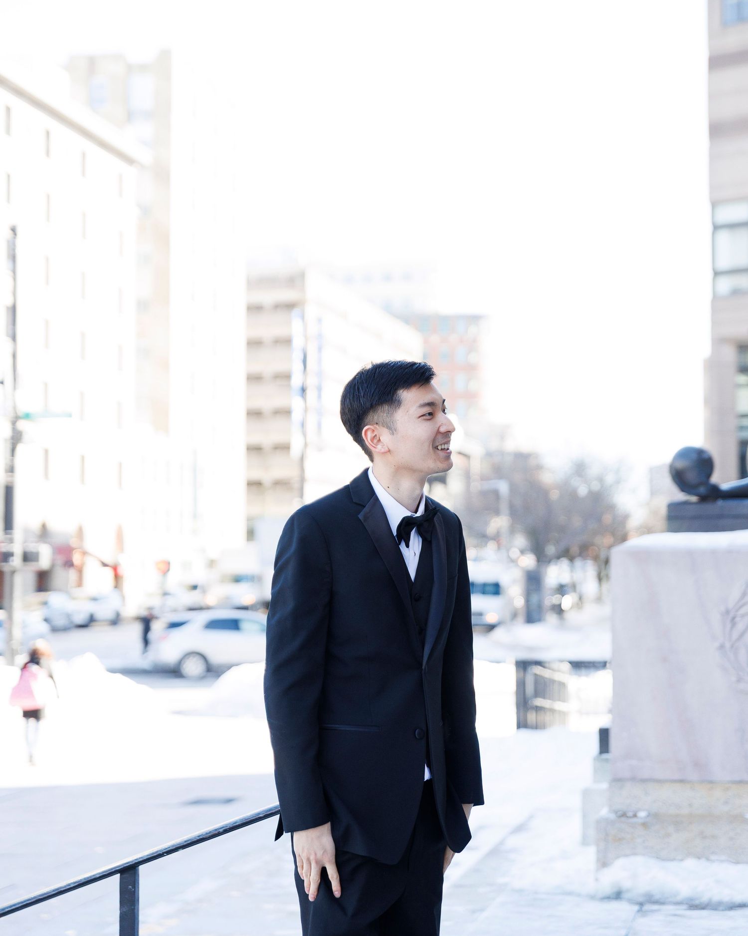 A person in a black coat laughs while standing in a snowy winter setting near a white building.