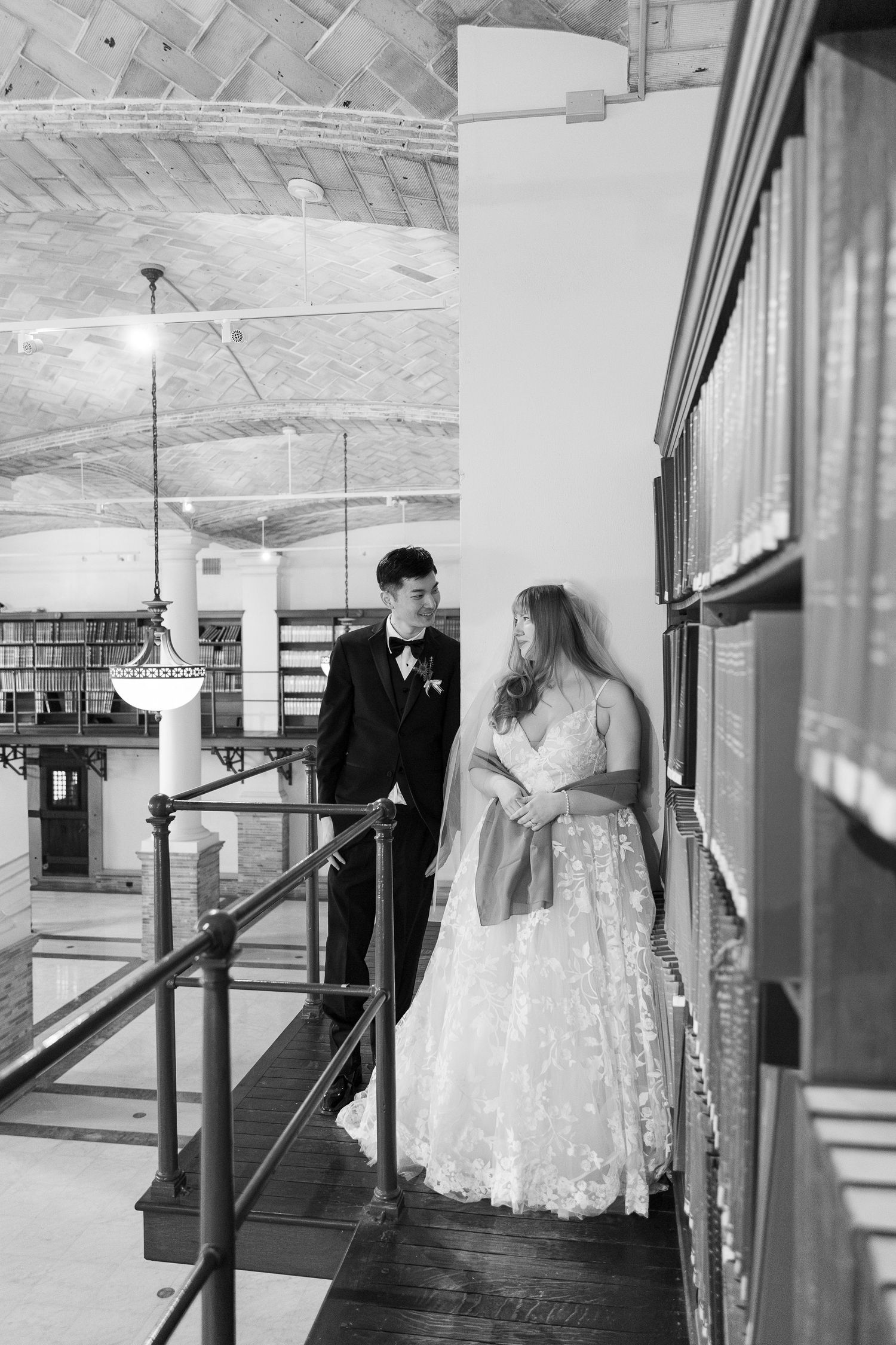 Black and white wedding photo of a couple standing on a balcony in a historic building with elegant chandeliers.