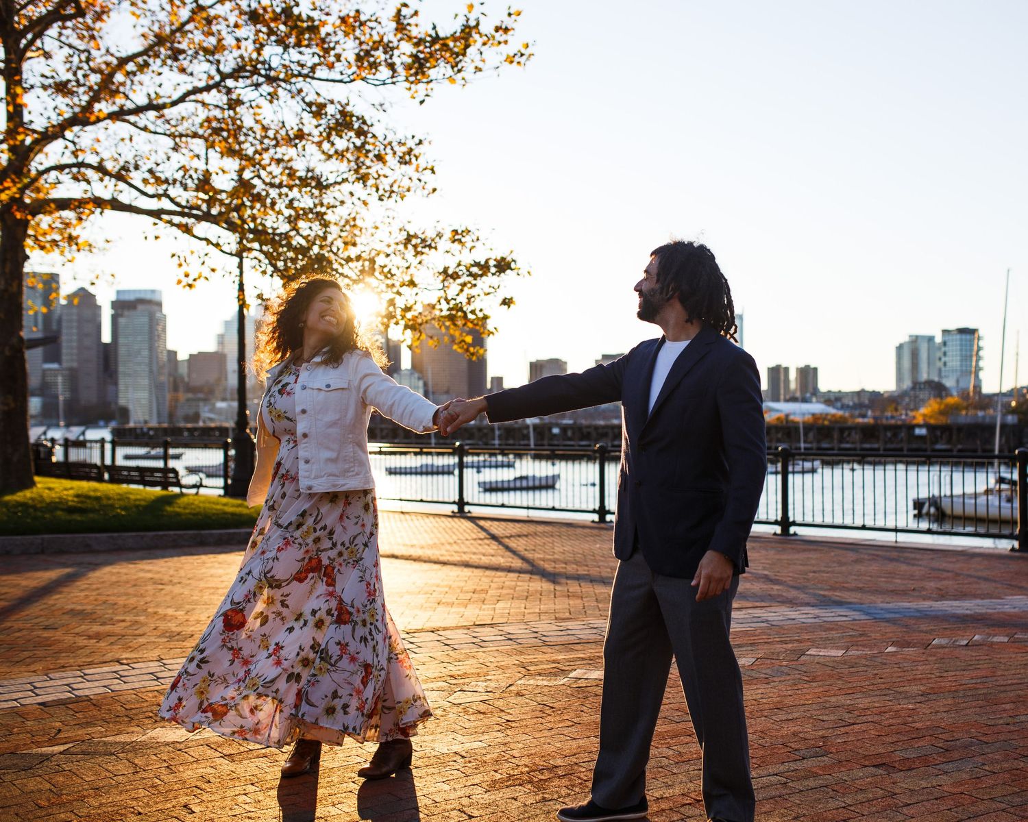 A romantic sunset engagement photoshoot along a brick waterfront promenade with autumn leaves and city skyline.