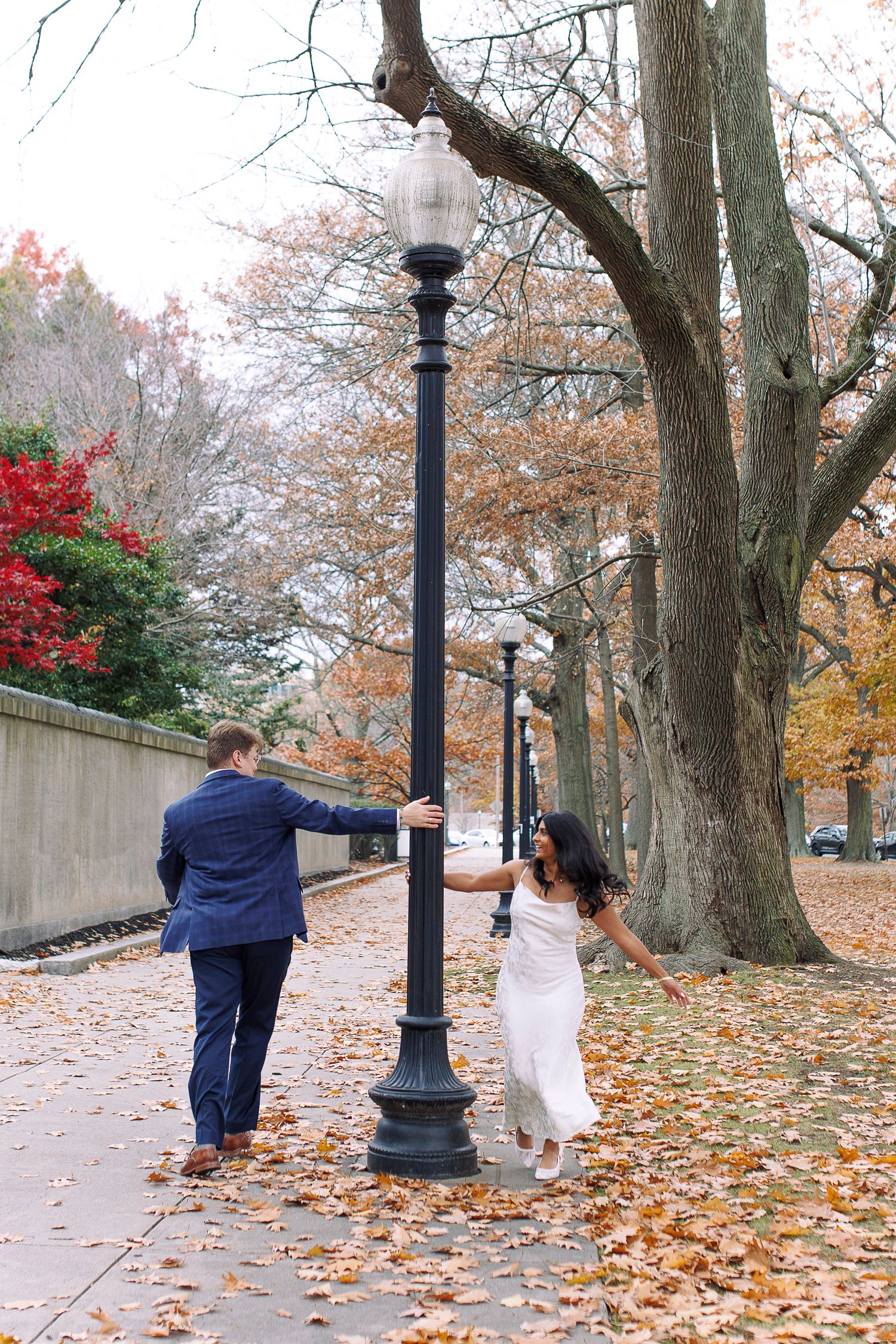 Two people walk together on a leaf-covered path through a park during autumn, with ornate lampposts lining the way.