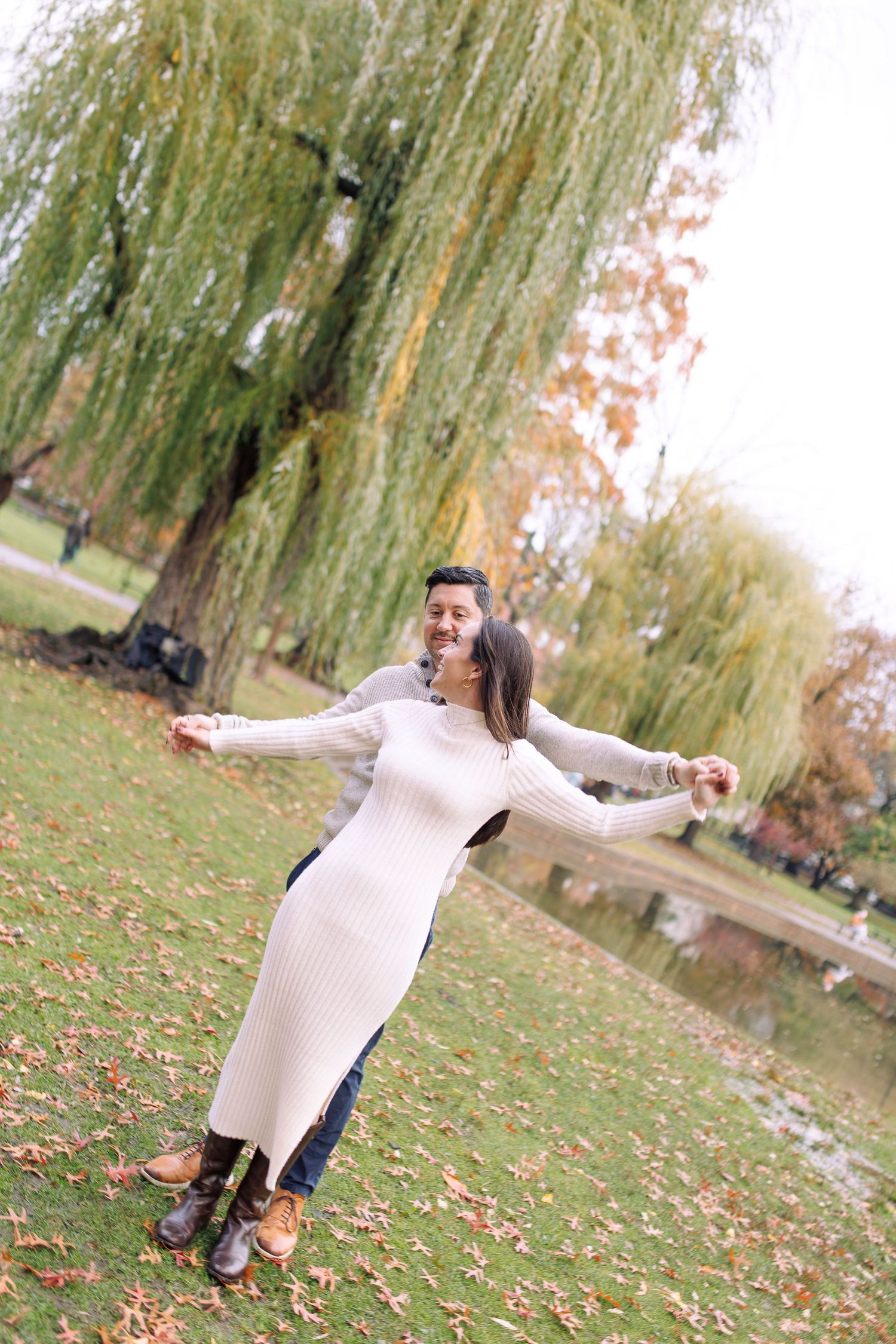 Someone in a beige sweater and dark pants twirls joyfully among autumn leaves beneath a weeping willow tree.