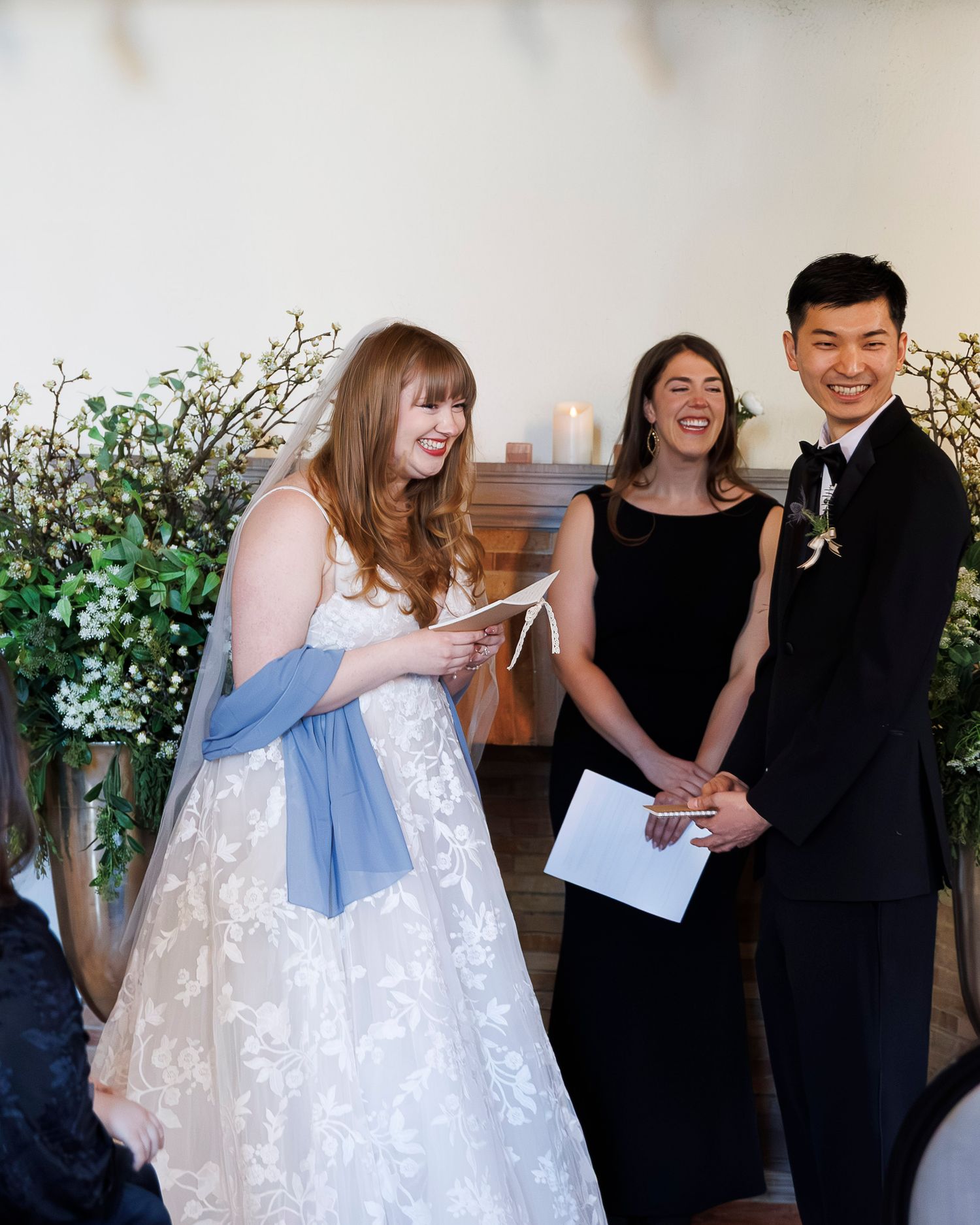 Wedding party gathered in formal attire with white floral decor and greenery in an indoor venue setting.