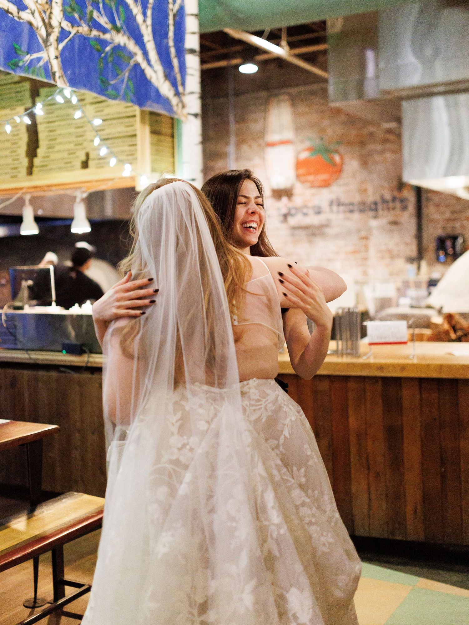 A bride in a white wedding dress laughs joyfully while sitting at a rustic bar counter in a restaurant.