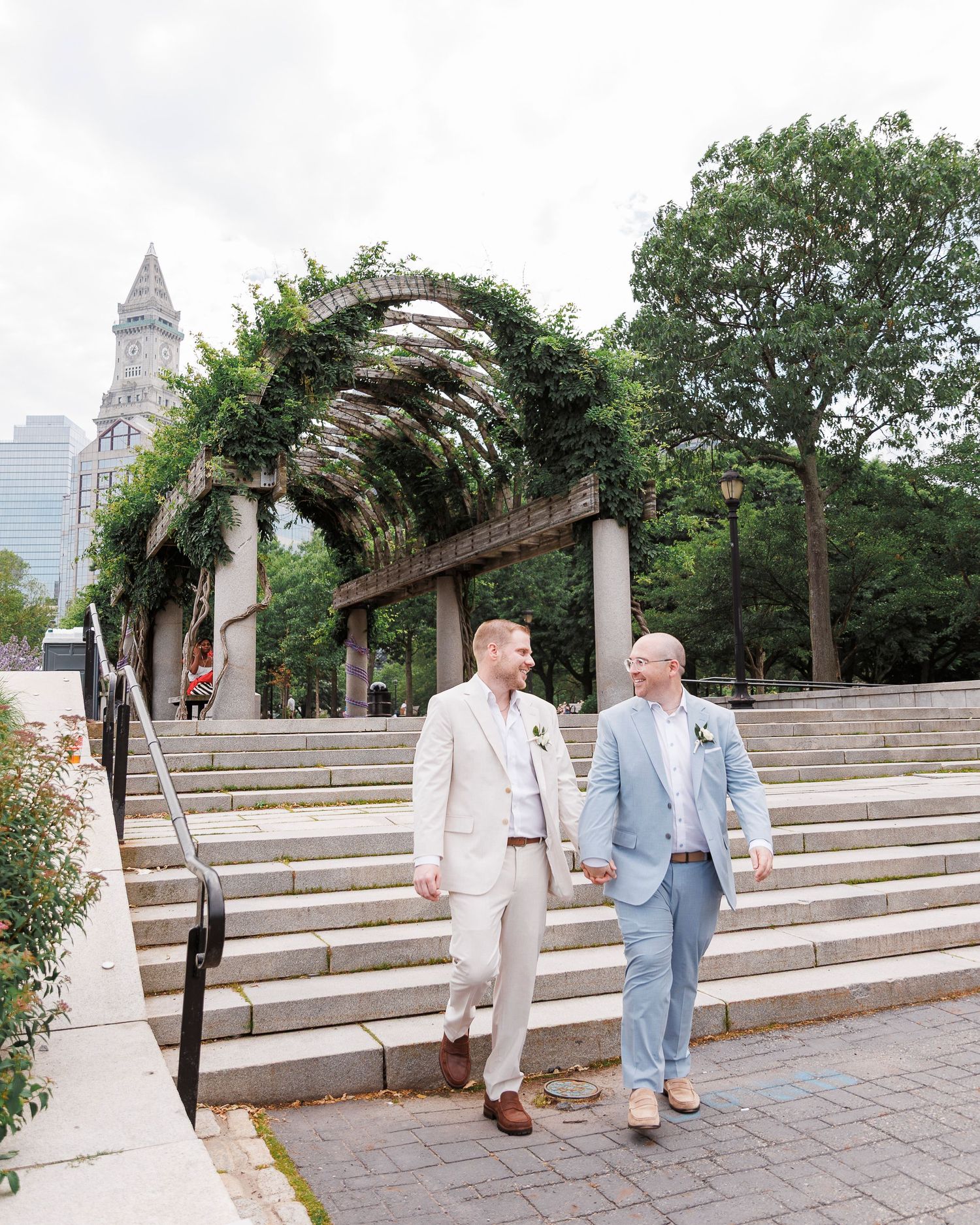 Two grooms hold hands walking near ivy-covered columns and stone steps at an outdoor wedding venue.