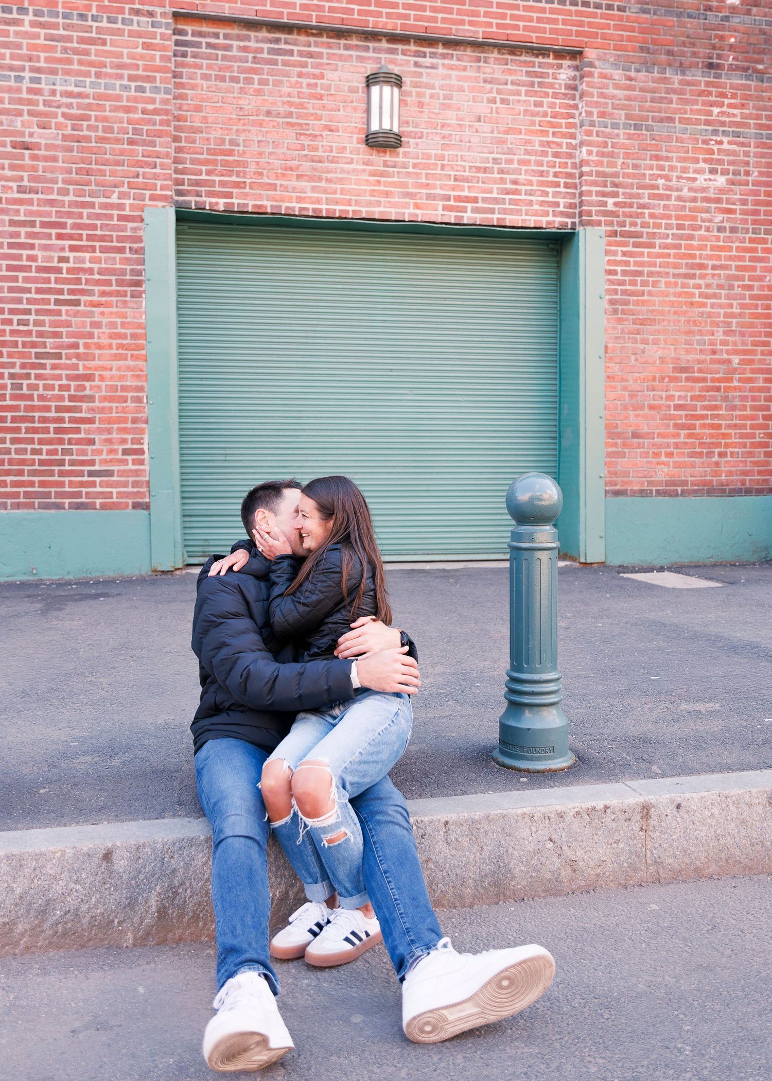 Couple sharing a romantic kiss while sitting on a curb in front of a brick wall with a green garage door.