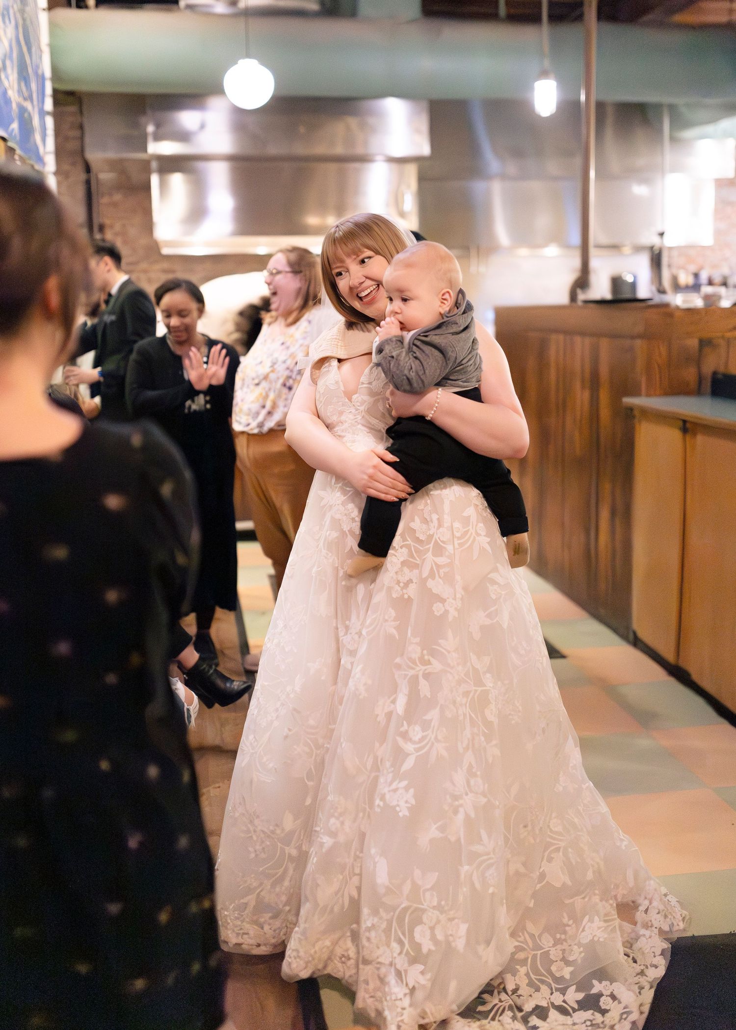 A touching moment at a wedding reception shows a bride in a lace gown holding a small baby while others look on.