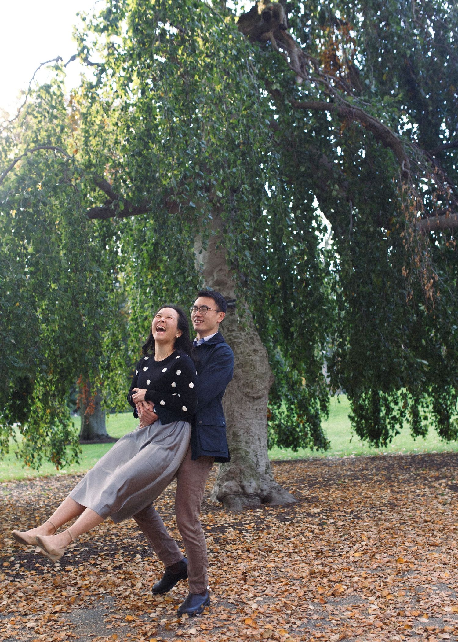 A couple sits and laughs together under a weeping willow tree on a carpet of fallen autumn leaves.