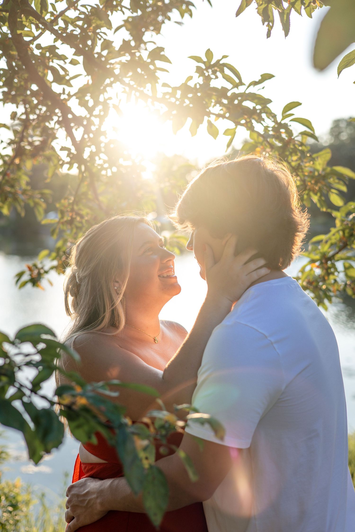 Joyful couple embracing and laughing under sunlit tree branches near a lake at golden hour.