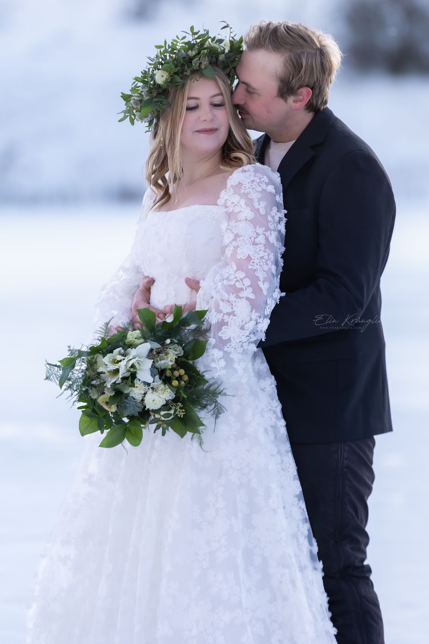 Winter wedding couple embraces in the snow with bride wearing lace dress and holding greenery bouquet.