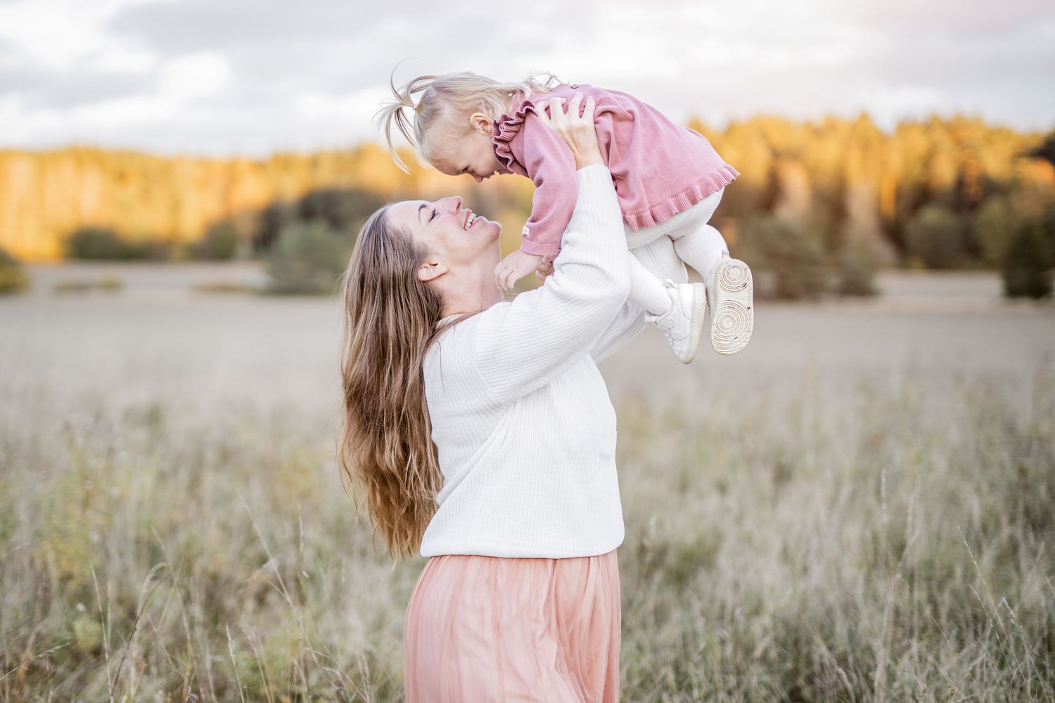 Mother and child embrace in a sunlit field wearing coordinated pink and white outfits.