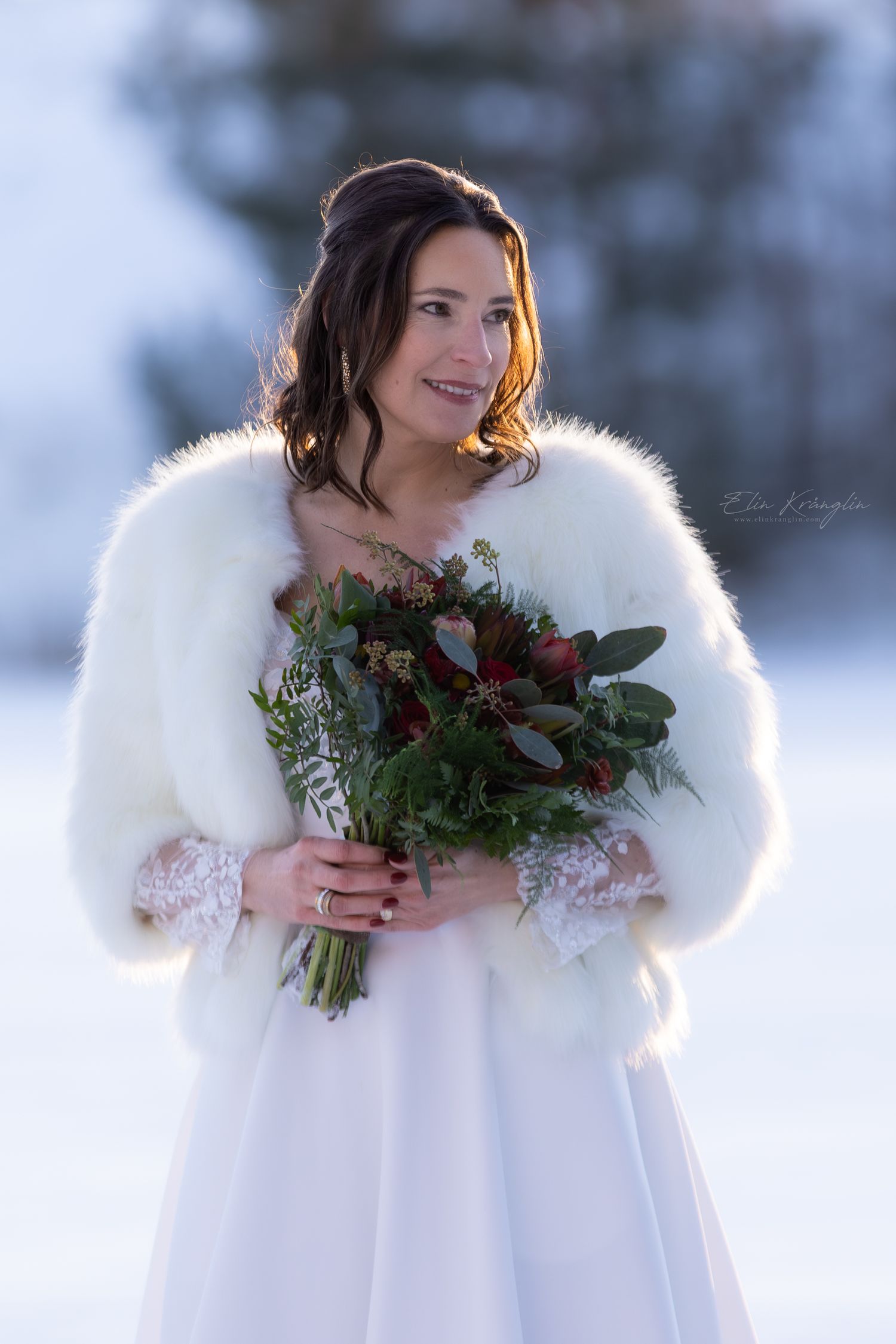 Winter bride in white fur wrap holds festive red and green bouquet against snowy backdrop.