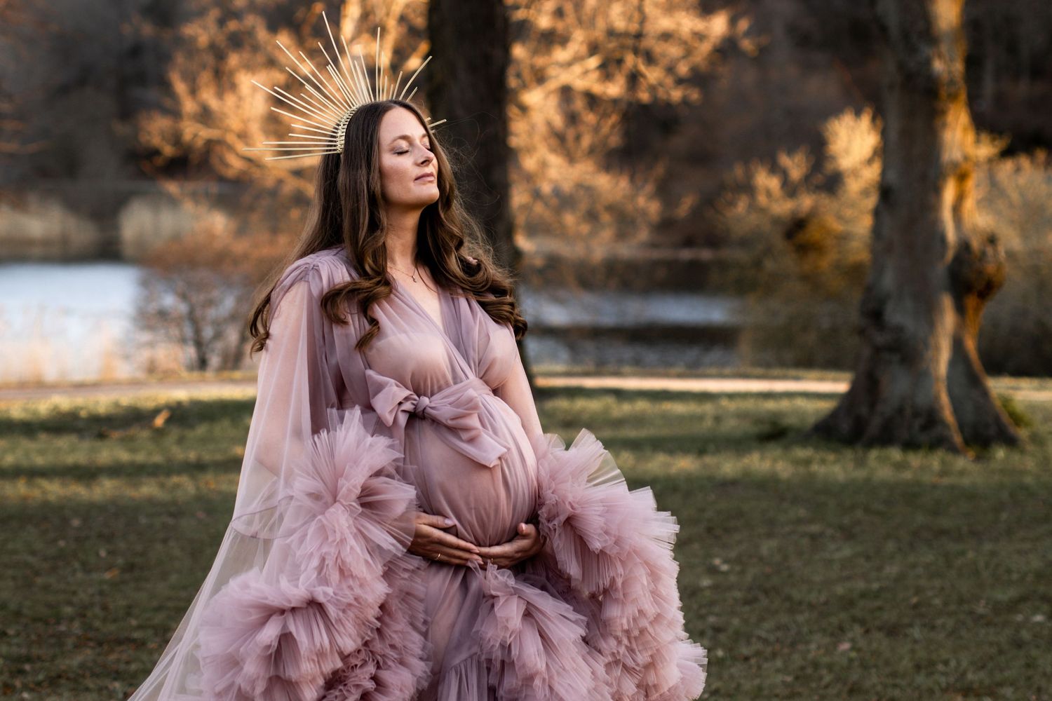 Pregnant woman in pink tulle gown with gold crown poses outdoors in autumn park during golden hour maternity photoshoot.