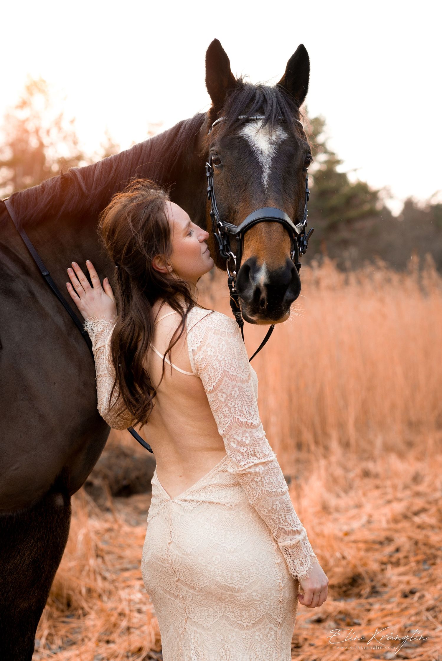 Woman in lace dress poses with dark horse in sunlit field at golden hour.