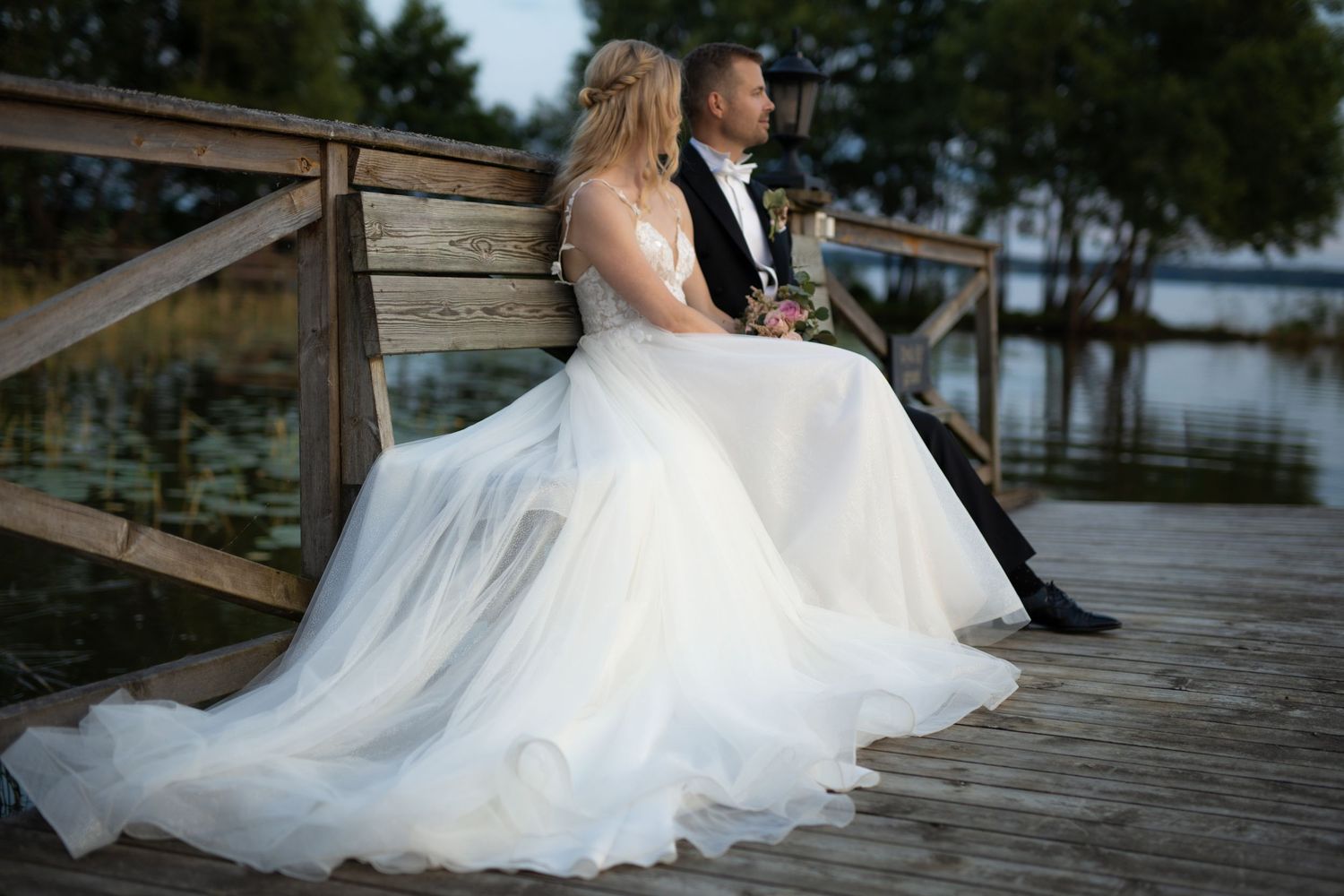 Couple sits on wooden dock with flowing white wedding dress.