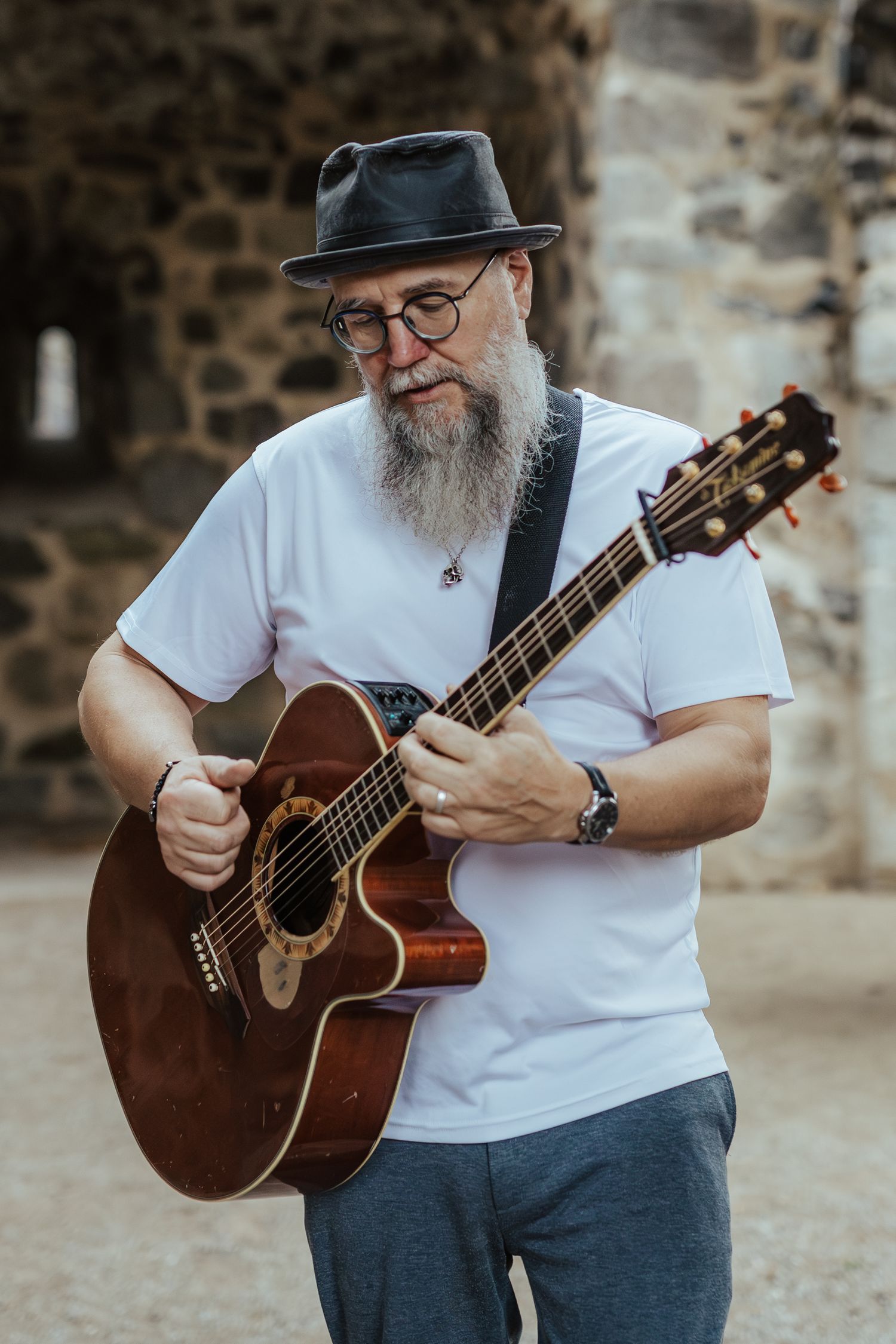 Musician in a white t-shirt and hat plays acoustic guitar against stone wall backdrop.