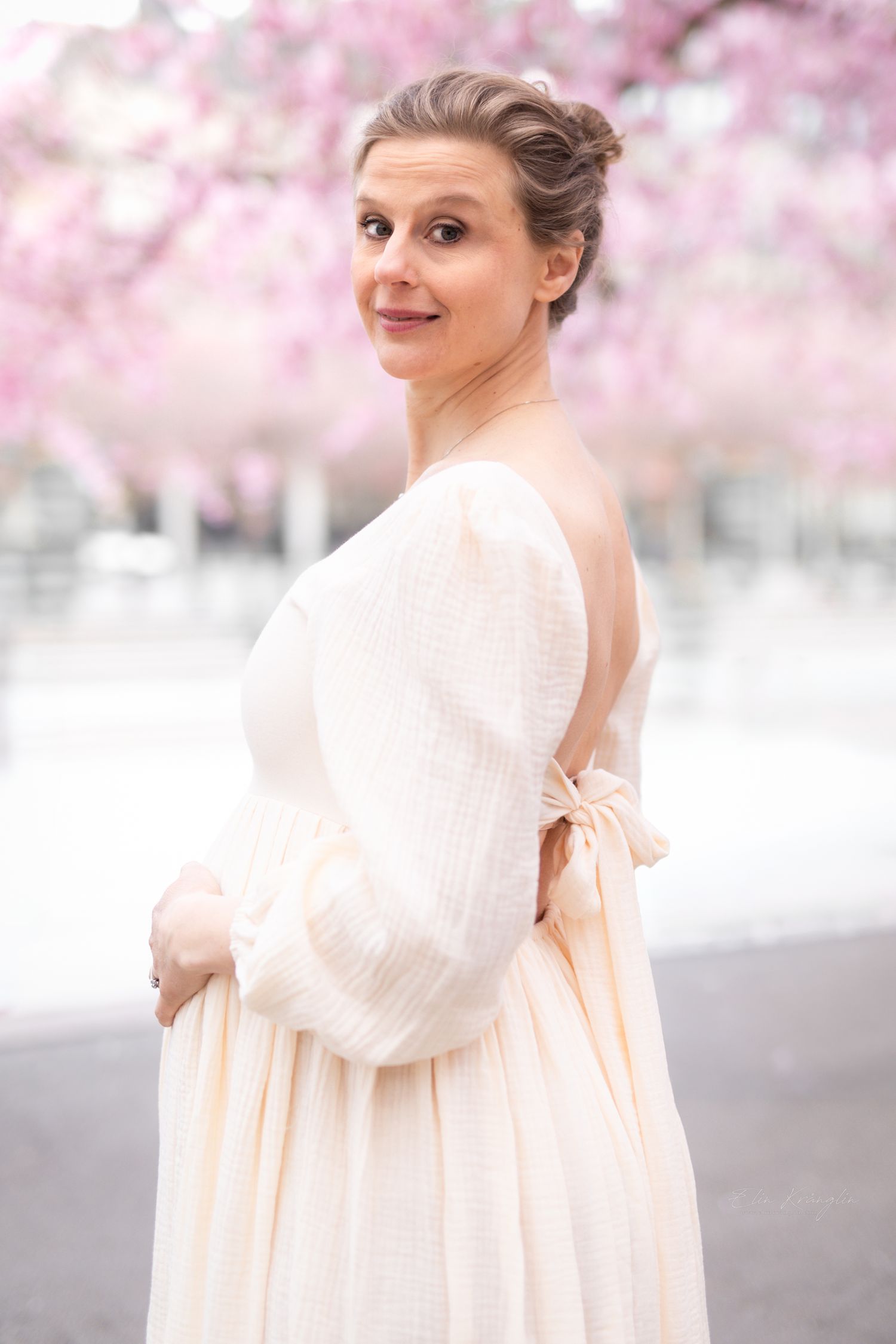 Pregnant woman in elegant backless cream dress poses gracefully among pink cherry blossoms in soft spring light.