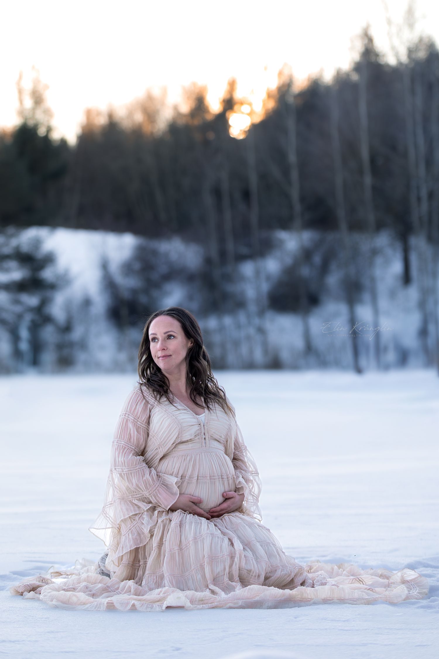 Maternity session features cream-colored dress against snowy winter landscape.
