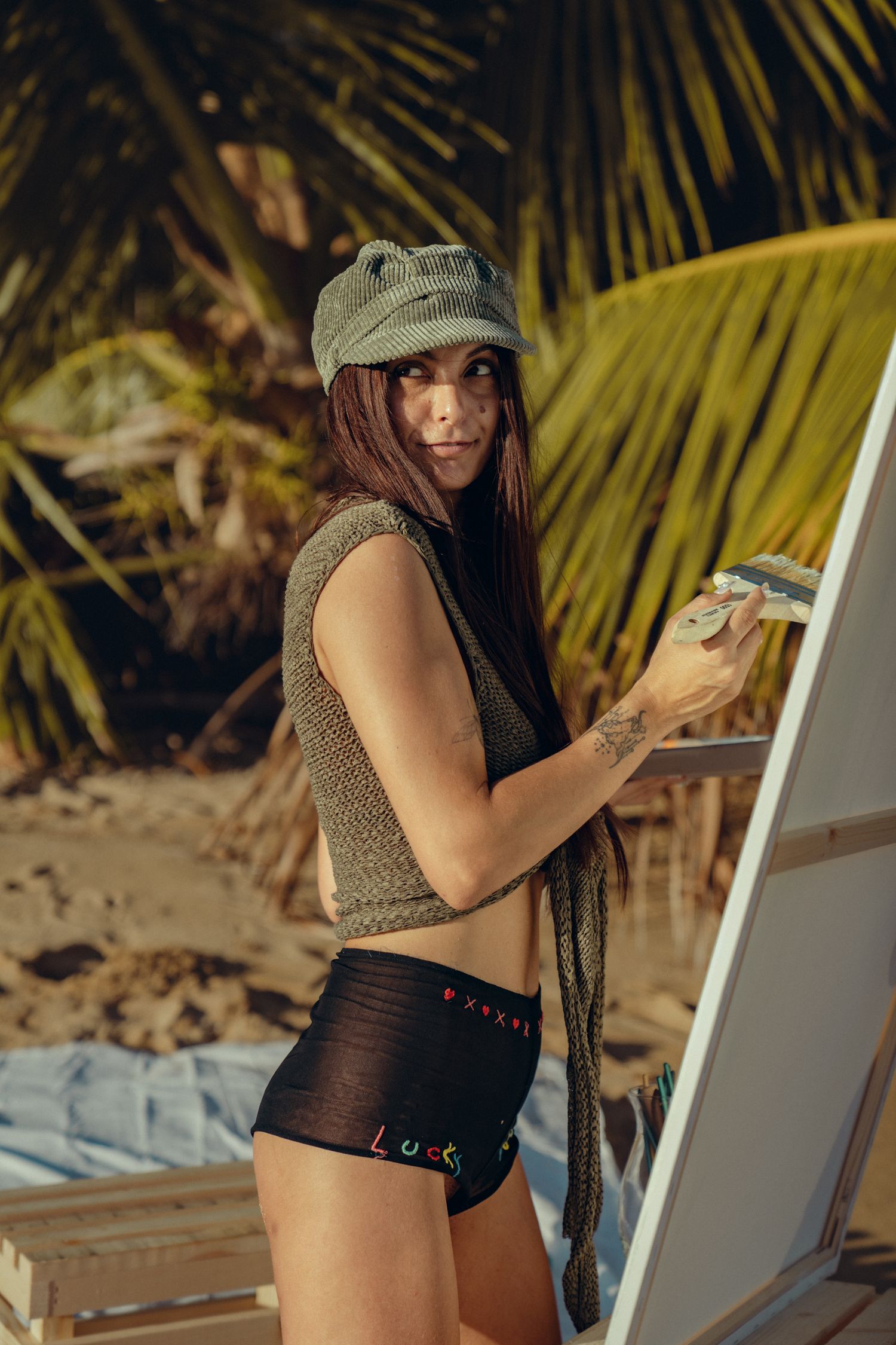 Person in casual beach attire and hat poses on sandy beach with palm trees in background.
