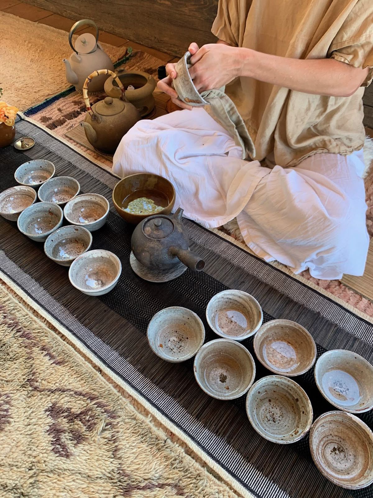 Person in linen clothing performing a traditional tea ceremony with rustic ceramic cups on a woven mat.