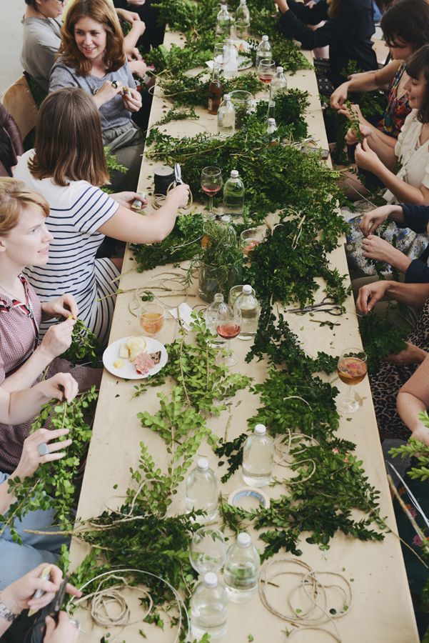 Group of women crafting floral wreaths with fresh greenery at a long wooden table during a wreath-making workshop.