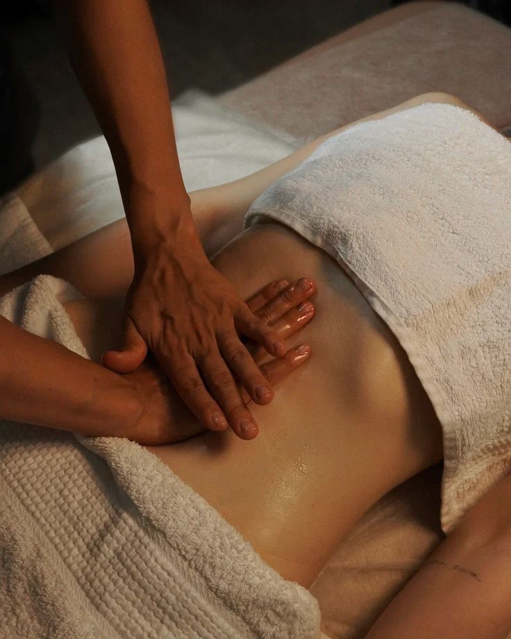 A massage therapist applies oil and performs a relaxing abdominal massage on a client draped in white towels.