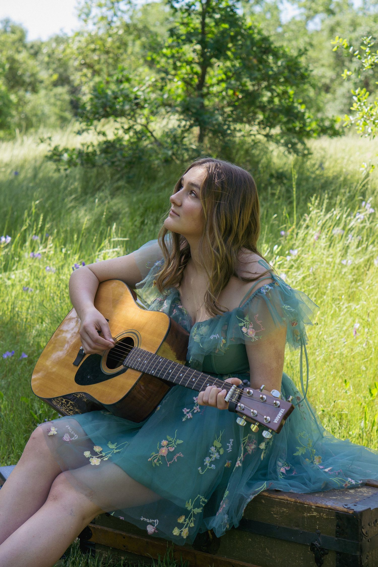 Someone relaxes in a turquoise dress with an acoustic guitar amidst a lush green field.