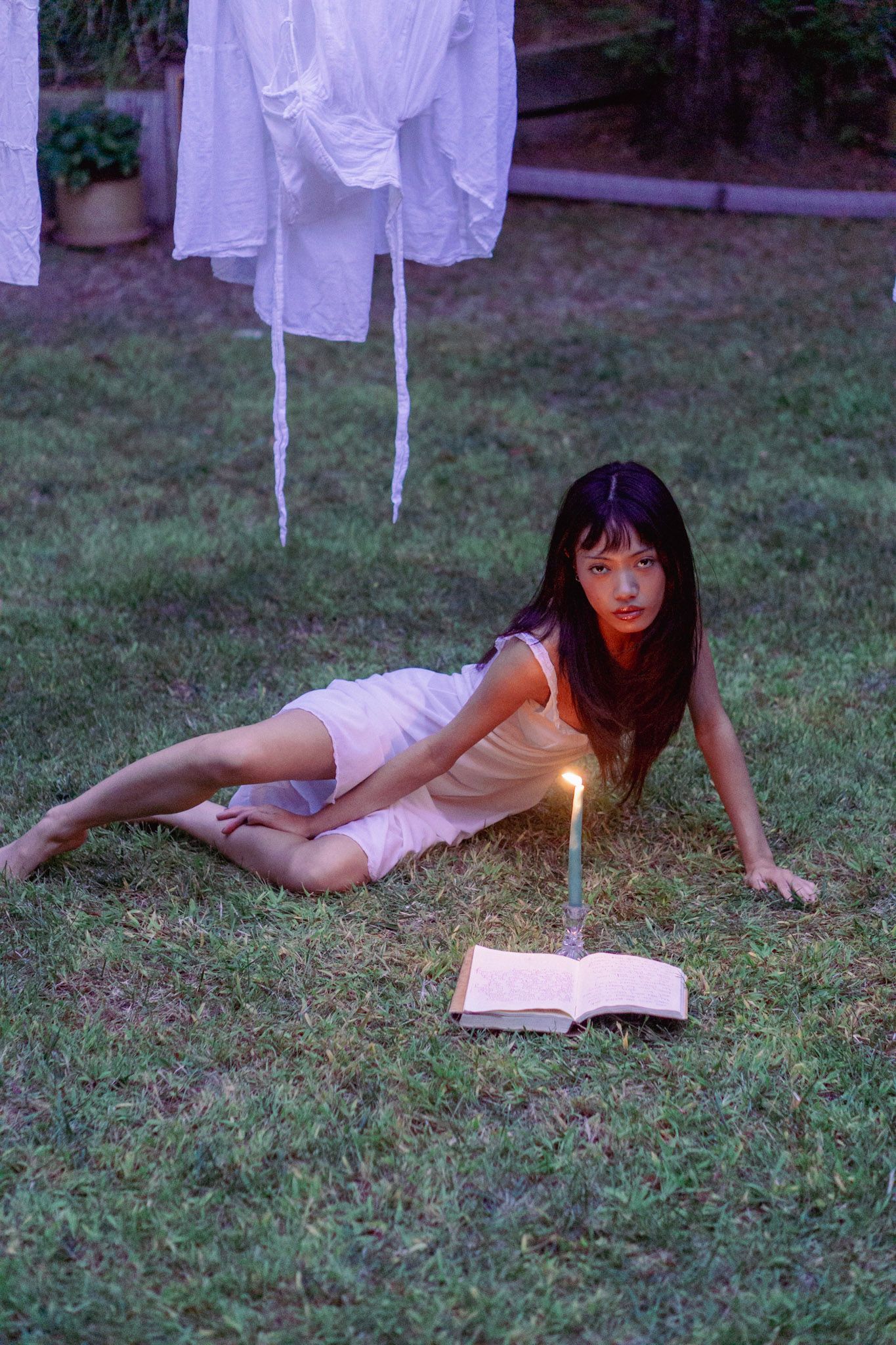 Person lying on grass reading book by candlelight with white linens hanging.