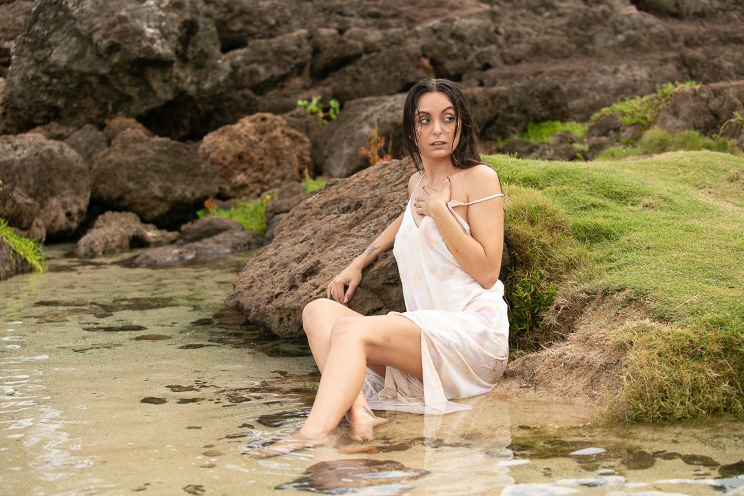Woman in white dress relaxes on rocky coastal landscape.