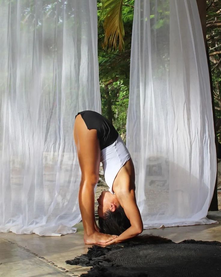 Woman performing a deep forward fold yoga pose outdoors near sheer white curtains and tropical greenery.