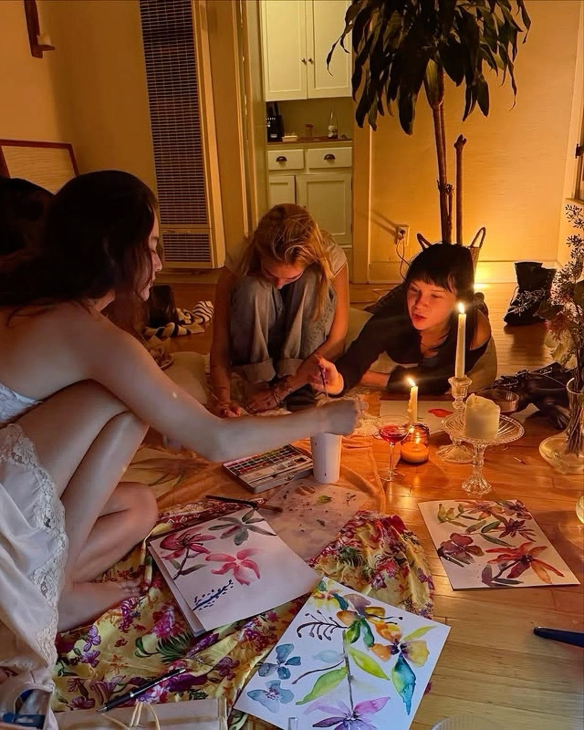 Three women sit on a floor by candlelight, painting colorful floral artwork on papers spread around them.