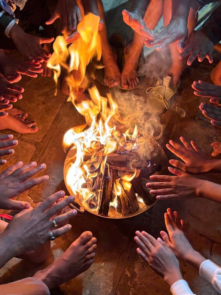 Group of people warming their hands around a glowing campfire in a metal bowl, flames rising with wooden logs.