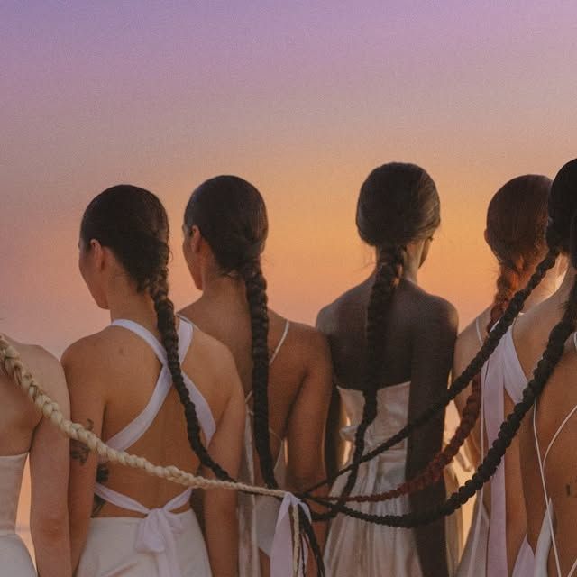 Four silhouettes with long braided hair stand against a sunset sky in matching white outfits.
