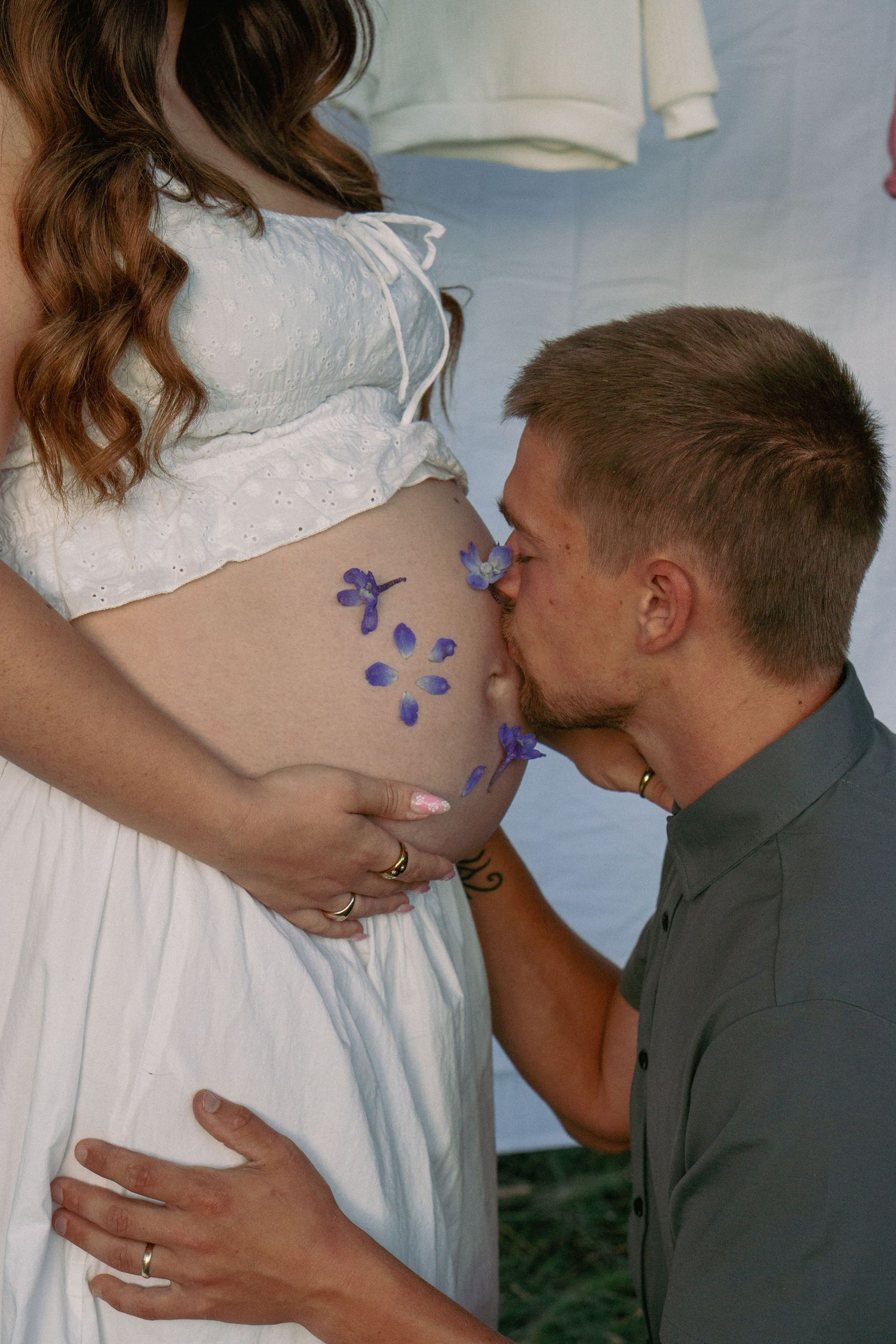 Man kissing pregnant woman's belly decorated with purple flowers during a maternity photo shoot outdoors.