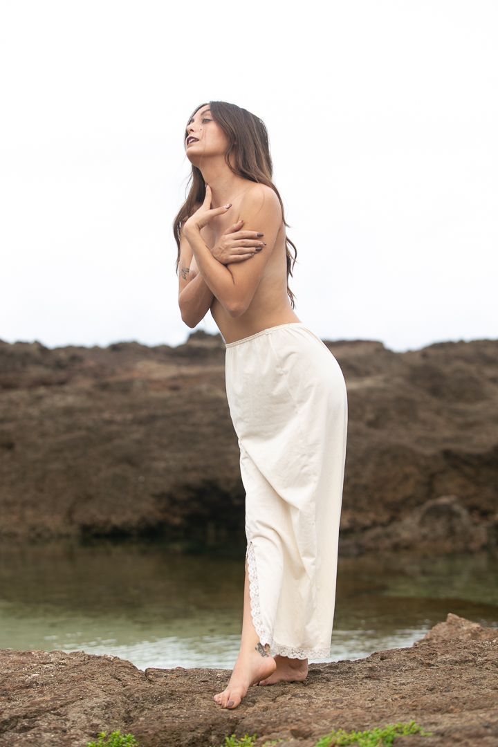 Woman in flowing white dress poses gracefully against rocky coastal backdrop.