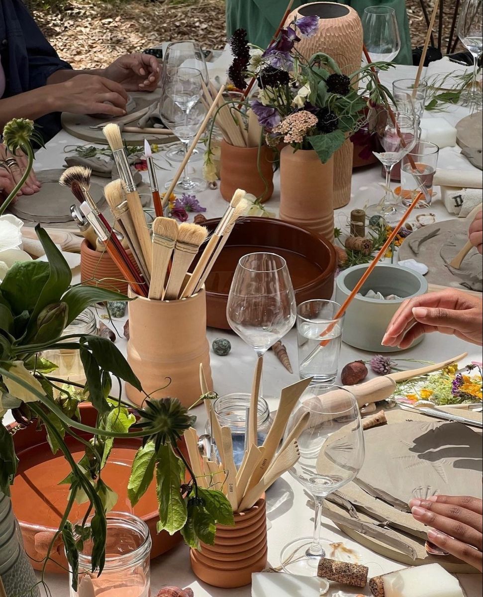 Outdoor art table setting with terracotta pots holding paintbrushes, wine glasses, floral arrangements, and palette plates.