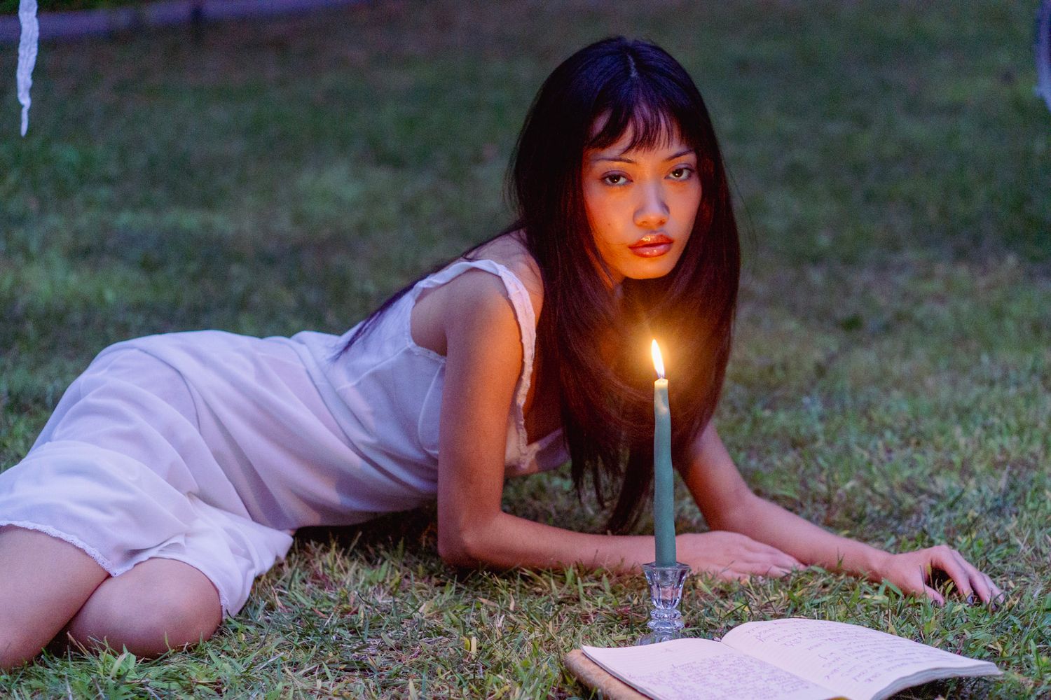 Person in white dress reading by candlelight on lawn.
