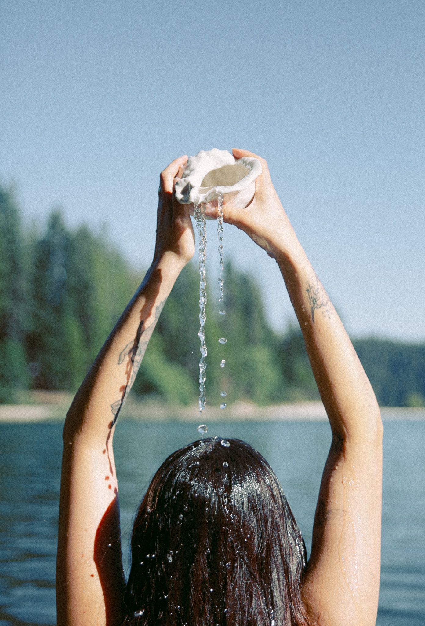 Water droplets falling through raised hands at lake with pine trees in background.