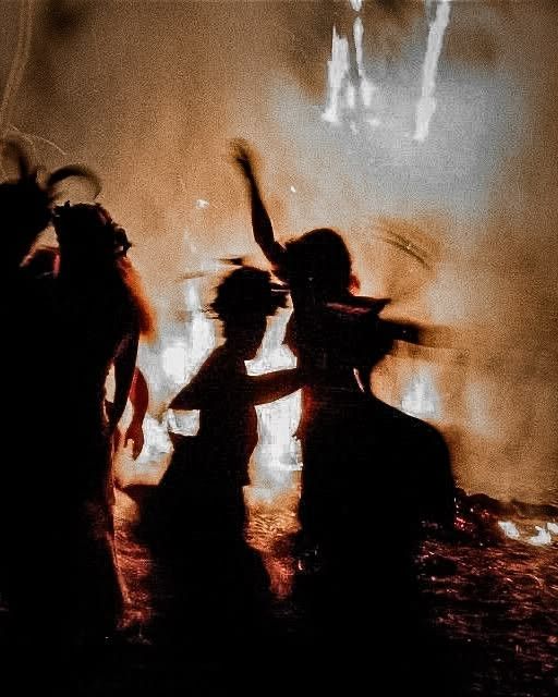 Silhouettes of people dancing around a large bonfire at night, with dramatic flames illuminating the scene.