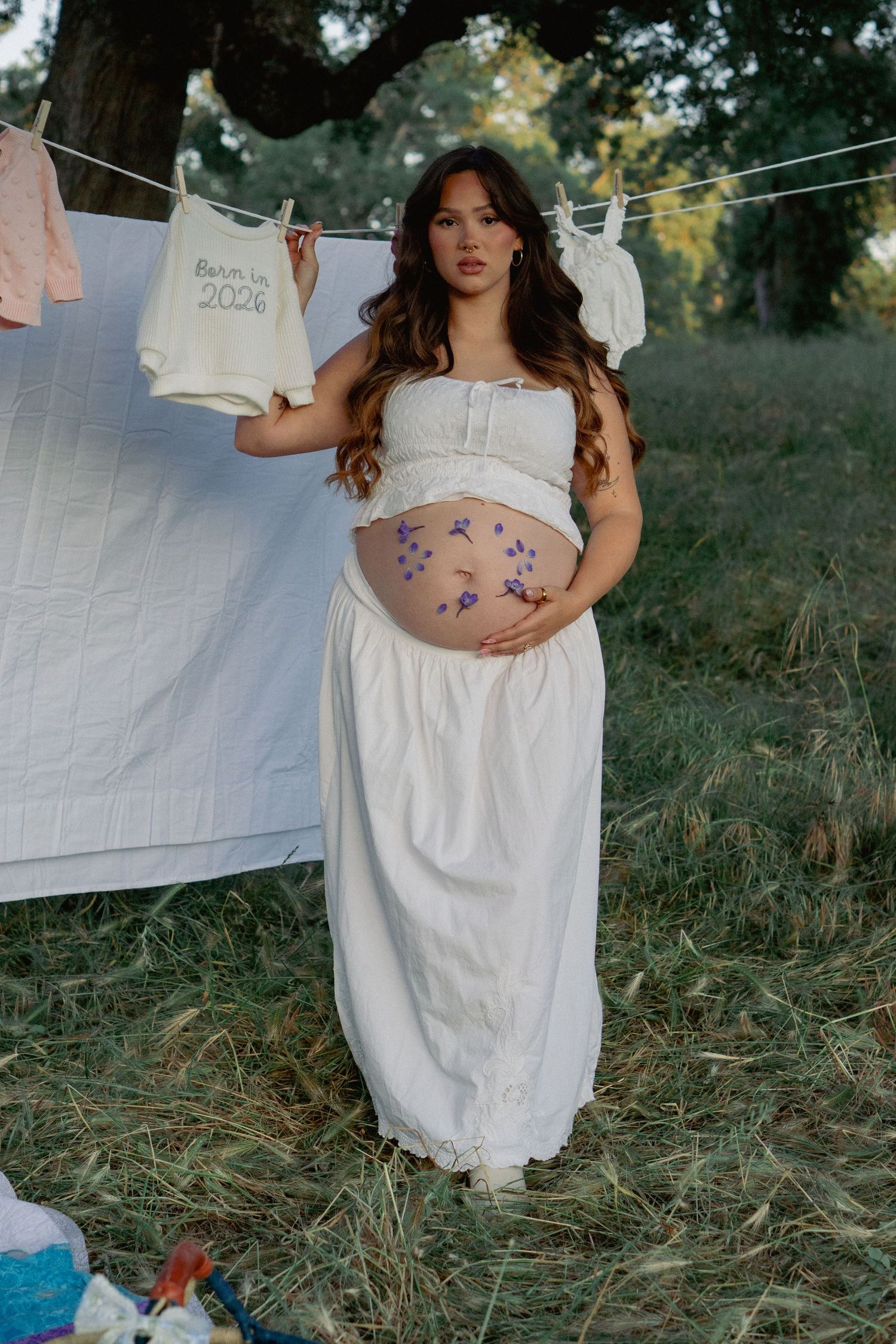 Pregnant woman in white crop top and skirt holds baby onesie outdoors near clothesline with purple flowers on belly.