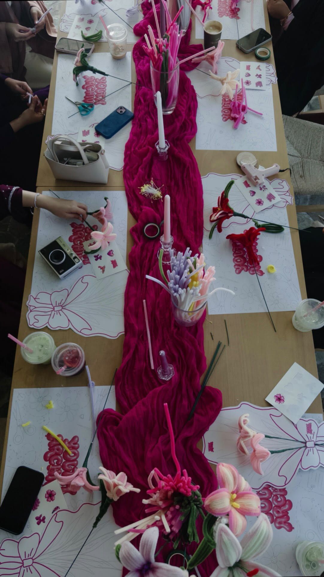 Overhead view of a pink floral craft table with a magenta runner, candles, flowers, and creative supplies.
