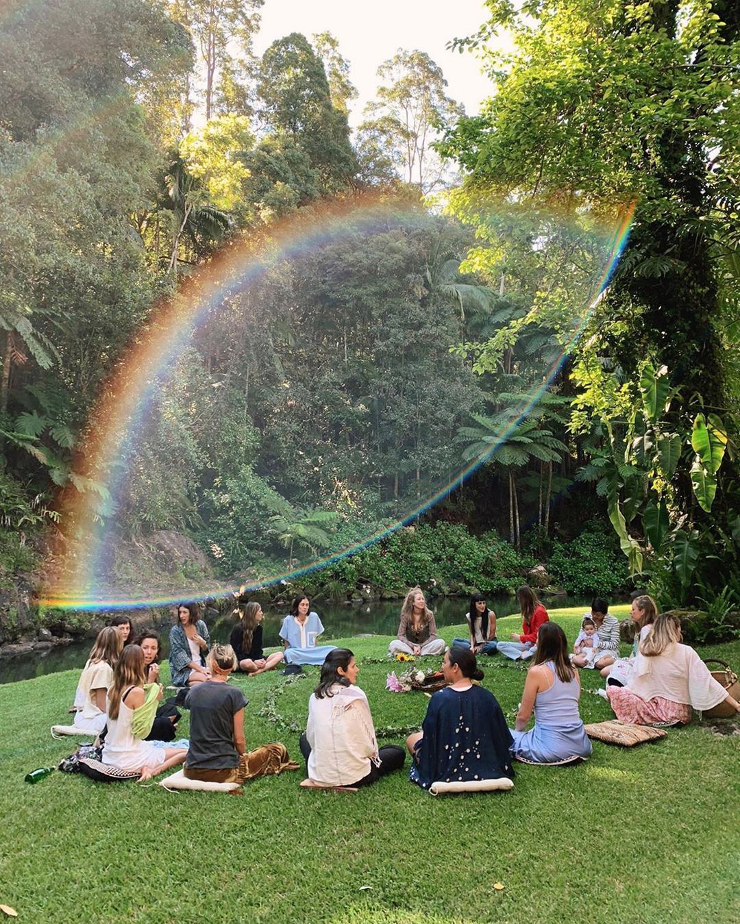 Group meditating on grass in lush green forest with a beautiful rainbow arching overhead during golden hour.