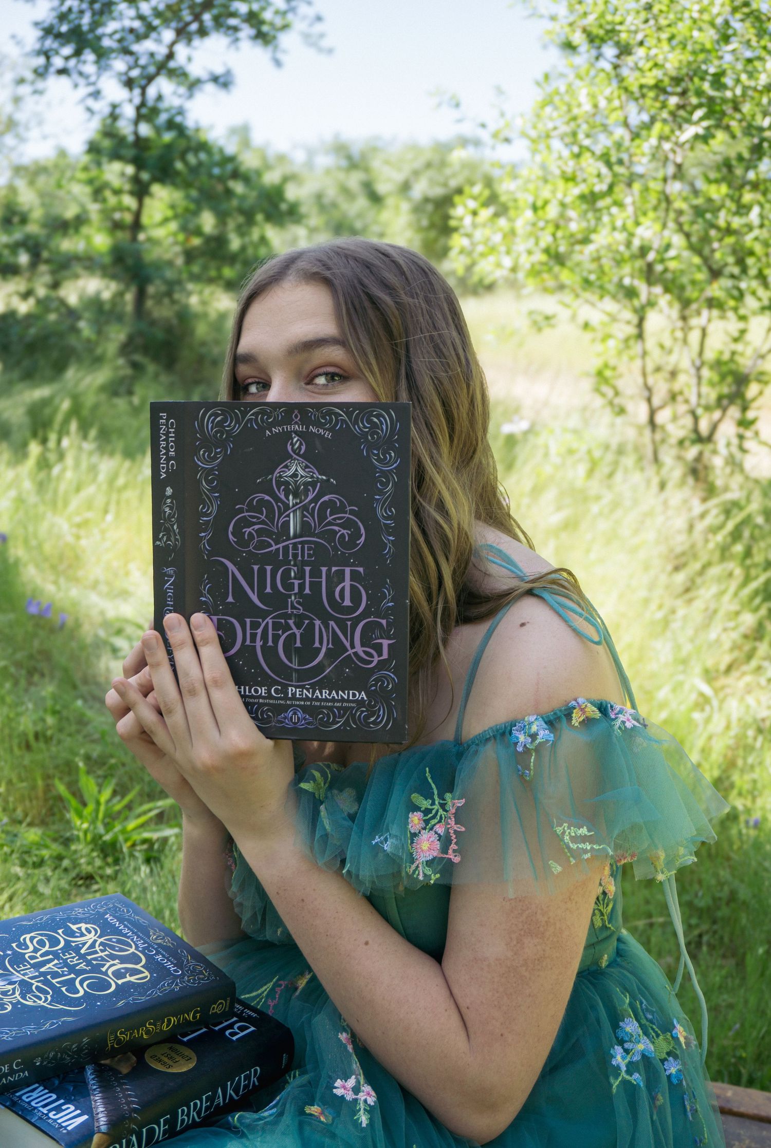 A turquoise dress-clad figure poses with a decorative book in a sunny meadow setting.