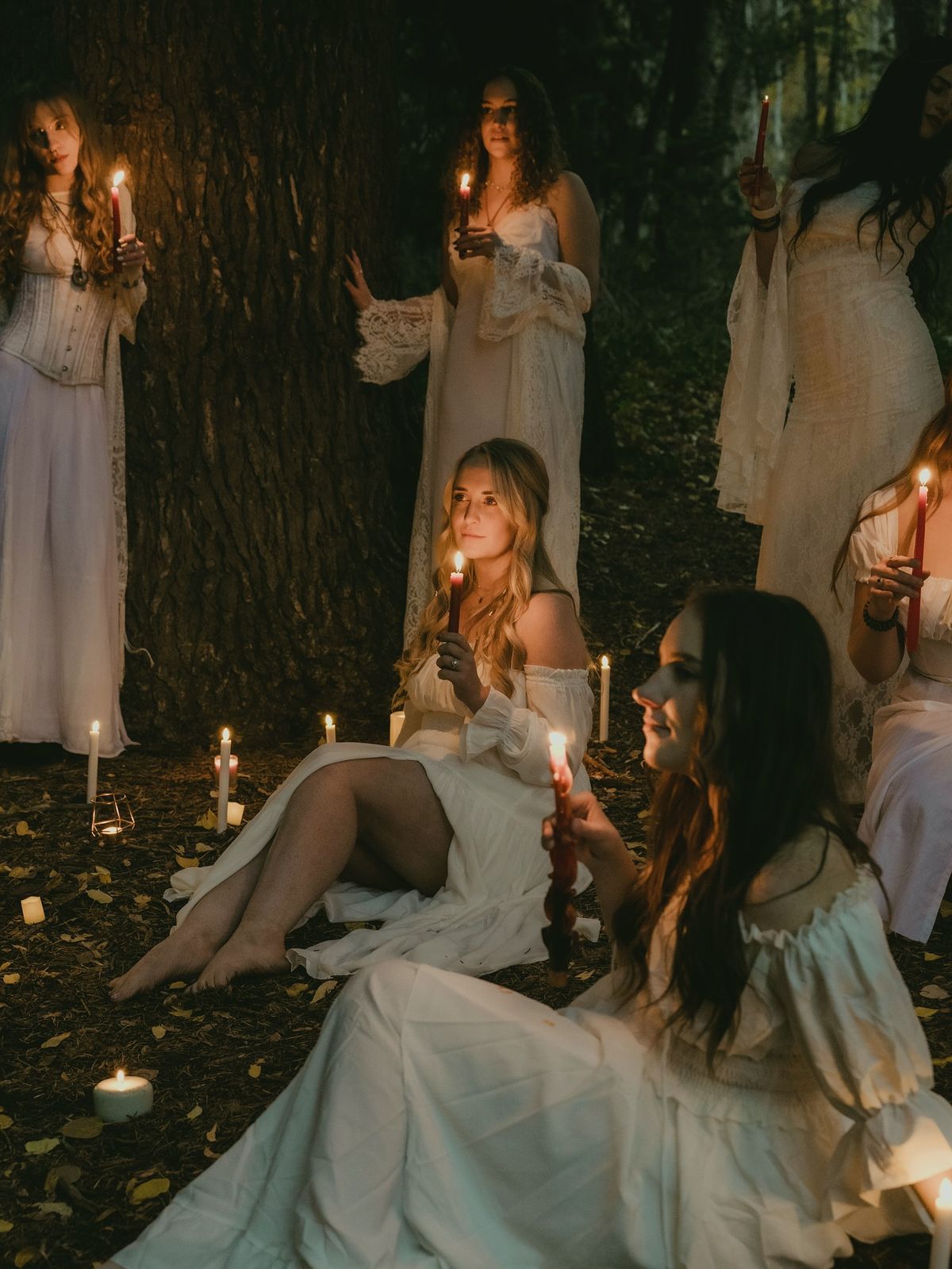 Women in white dresses holding candles gather in a mystical forest ritual at night.